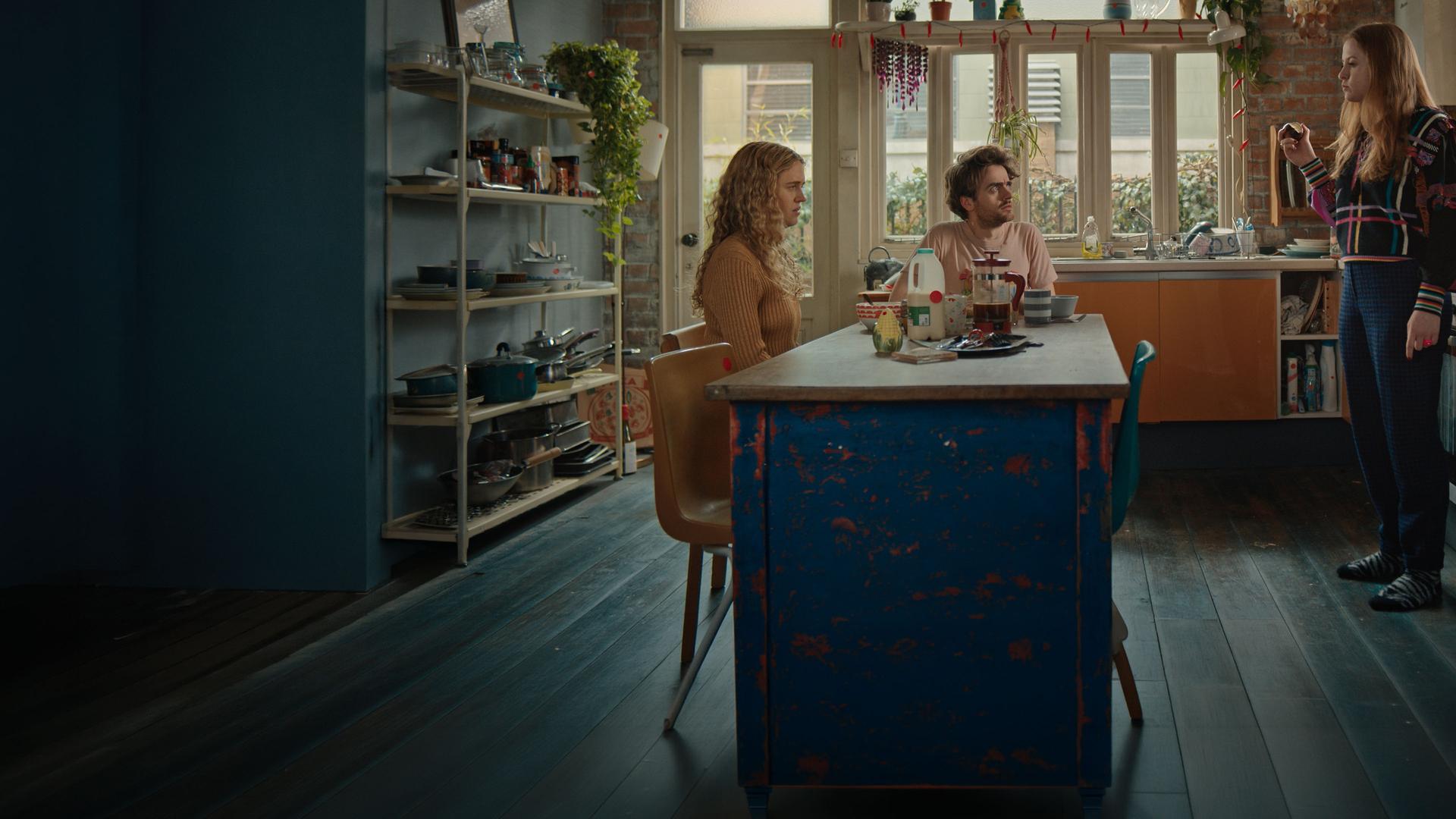 "Extraordinary - Unsere Freunde sind die wahren Superkräfte": Carrie (Sofia Oxenham), Jizzlord (Luke Rollason) und Jen (Máiréad Tyers) sind gemeinsam in der Küche. Jen steht, Carrie und Jizzlord sitzen am gedeckten Tisch. Jen beißt demonstrativ in ihre Zwiebel.