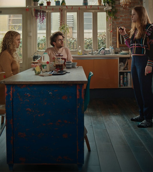 "Extraordinary - Unsere Freunde sind die wahren Superkräfte": Carrie (Sofia Oxenham), Jizzlord (Luke Rollason) und Jen (Máiréad Tyers) sind gemeinsam in der Küche. Jen steht, Carrie und Jizzlord sitzen am gedeckten Tisch. Jen beißt demonstrativ in ihre Zwiebel.