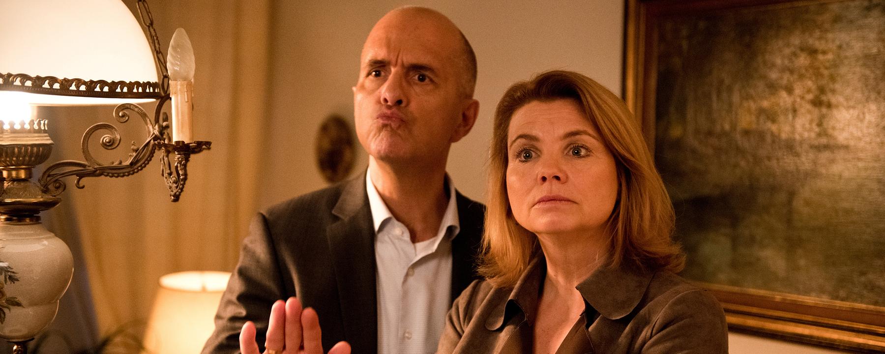 Erik Merz (Christoph Maria Herbst) und Anne Merz (Annette Frier) stehen in einem altmodischen und rustikal eingerichteten Zimmer nebeneinander. Auf Kopfhöhe hängt ein Lampenschirm von der Decke, der alles beleuchtet. Erik macht eine beschwichtigende Geste mit der Hand. Anne hat die Arme verschränkt und schaut mahnend.