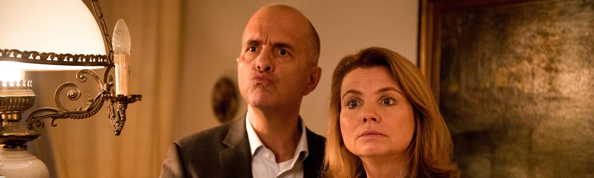 Erik Merz (Christoph Maria Herbst) und Anne Merz (Annette Frier) stehen in einem altmodischen und rustikal eingerichteten Zimmer nebeneinander. Auf Kopfhöhe hängt ein Lampenschirm von der Decke, der alles beleuchtet. Erik macht eine beschwichtigende Geste mit der Hand. Anne hat die Arme verschränkt und schaut mahnend.