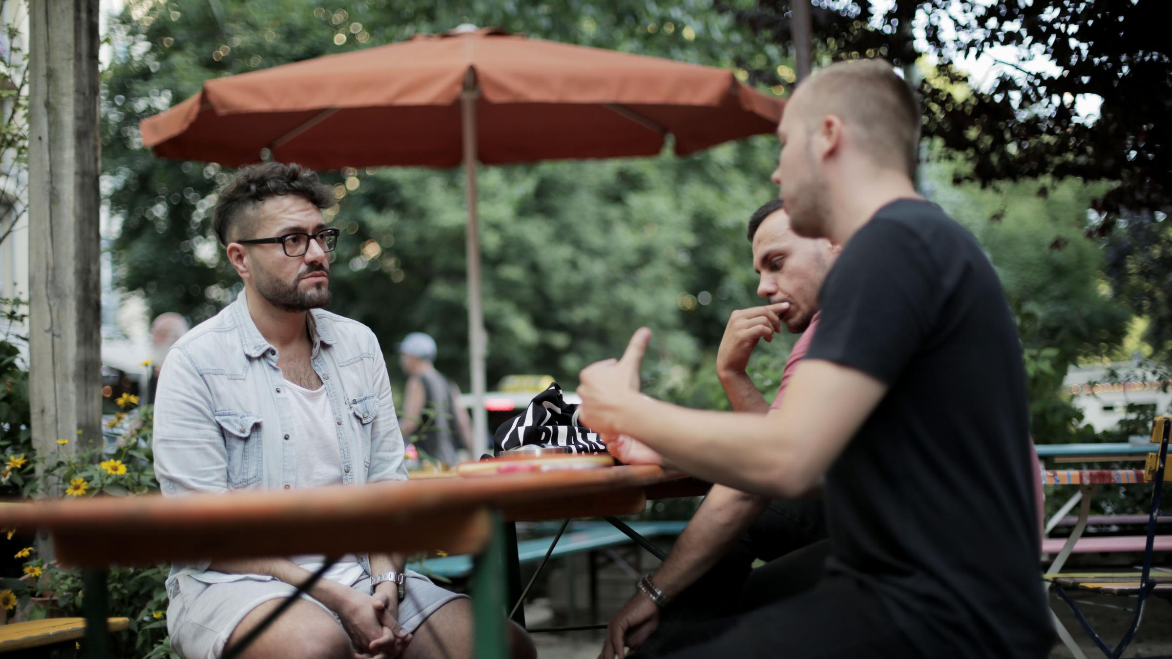 "Unter Beobachtung - Muslimische Männer in Deutschland": Ahmad Omeirate (links) im Gespräch mit Dominic Jóżwiak (vorne rechts) und Osan Yaran (hinten rechts). Sie sitzen im Freien an einem Biertisch.