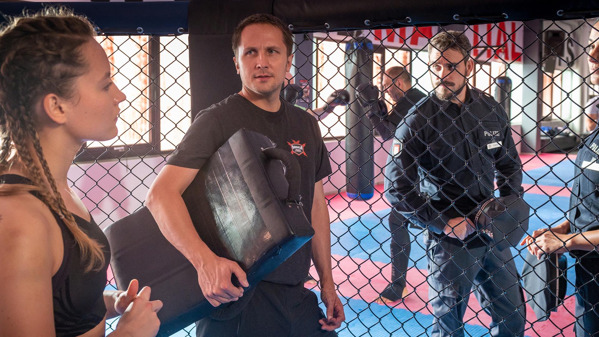 Die Kampfsportlerin Inga Sandmeier (Ella Morgen) steht in einem Kampfkäfig. Ihr Trainer Leon Hübner (Lasse Myhr) steht seitlich neben ihr und lehnt am Käfiggitter. Auf der anderen Seite befinden sich die Polizistin Pinar Aslan (Aybi Era) und ihr Kollege Kris Freiberg (Marc Barthel).