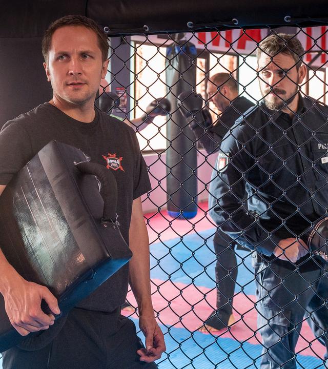 Die Kampfsportlerin Inga Sandmeier (Ella Morgen) steht in einem Kampfkäfig. Ihr Trainer Leon Hübner (Lasse Myhr) steht seitlich neben ihr und lehnt am Käfiggitter. Auf der anderen Seite befinden sich die Polizistin Pinar Aslan (Aybi Era) und ihr Kollege Kris Freiberg (Marc Barthel).