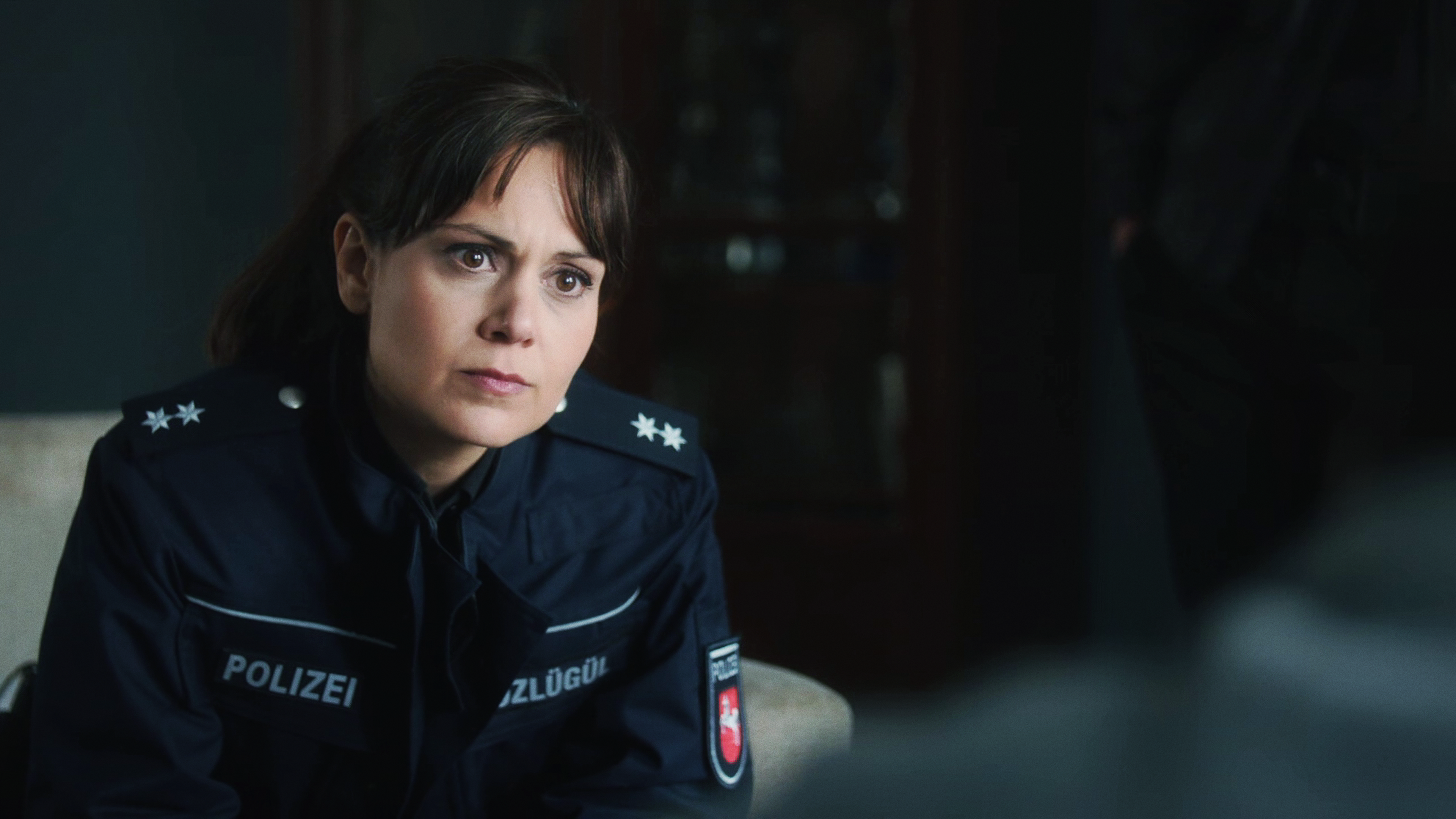 Süher Özlügül (Sophie Dal) sitzt auf einer Couch und spricht mit jemandem, der nicht zu erkennen ist. 