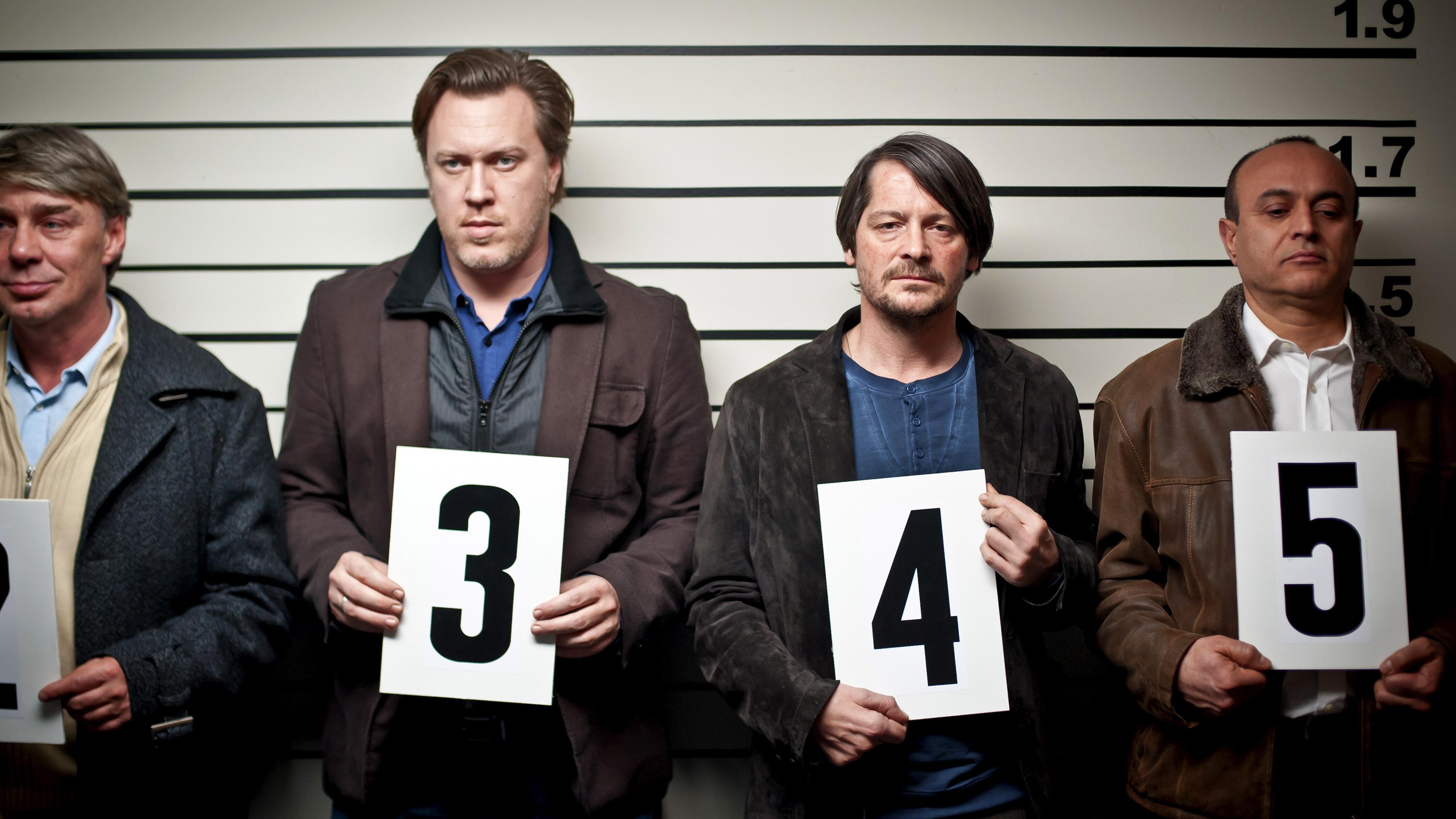 Mario Diller (Nicholas Ofczarek), Erich Kessel (Fritz Karl) und zwei weitere Männer stehen für eine Gegenüberstellung an einer Wand. Jeder von ihnen hält eine weiße Karte mit einer Zahl darauf vor sich.