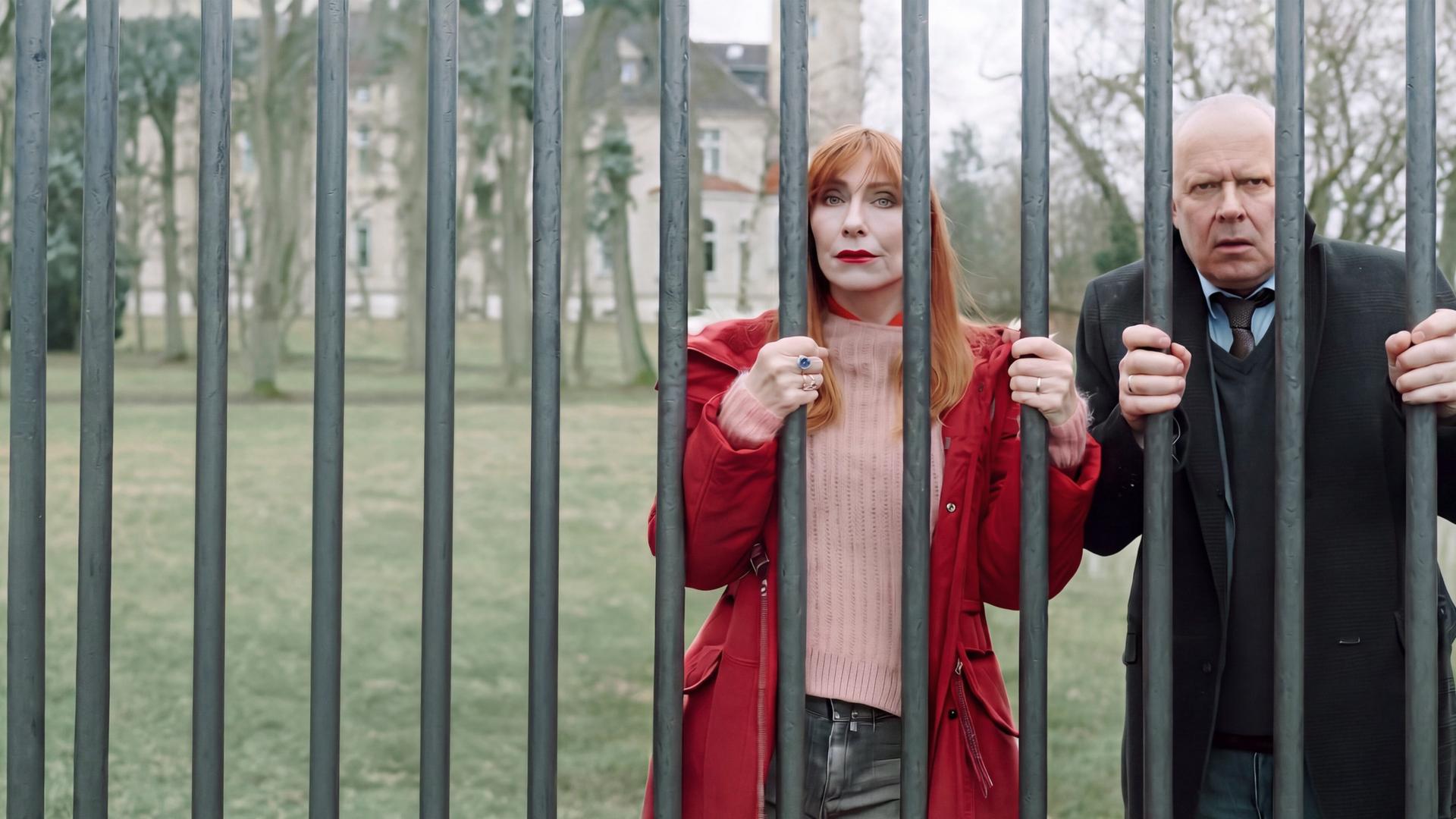 "Familie Bundschuh - Unter Verschluss": Gerald ( Axel Milberg) und Gundula (Andrea Sawatzki) krallen sich an ihrem Eisenzaun fest und blicken hilflos durch die dicken Gitterstäbe. Im Hintergrund ist der großangelegte Garten und ein Teil des Gutshauses zu sehen.