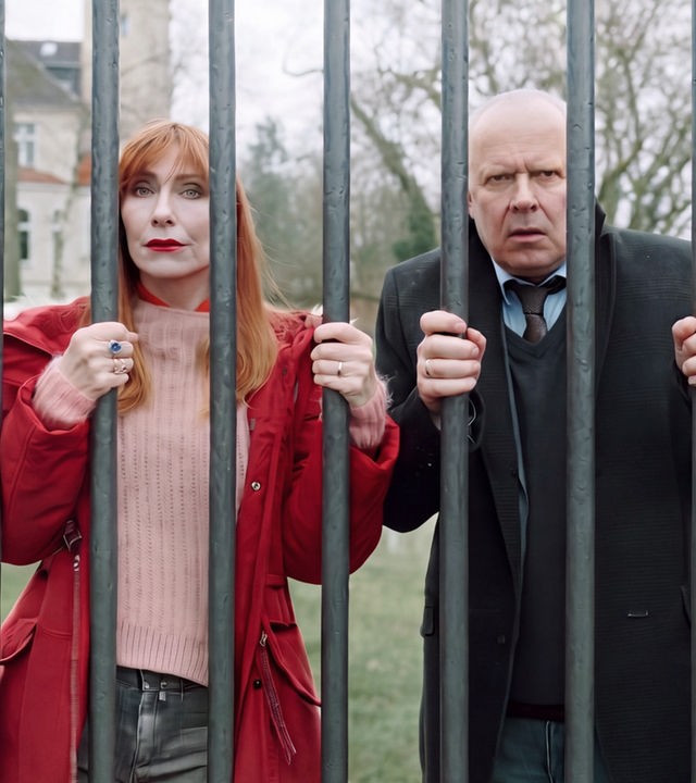 "Familie Bundschuh - Unter Verschluss": Gerald ( Axel Milberg) und Gundula (Andrea Sawatzki) krallen sich an ihrem Eisenzaun fest und blicken hilflos durch die dicken Gitterstäbe. Im Hintergrund ist der großangelegte Garten und ein Teil des Gutshauses zu sehen.