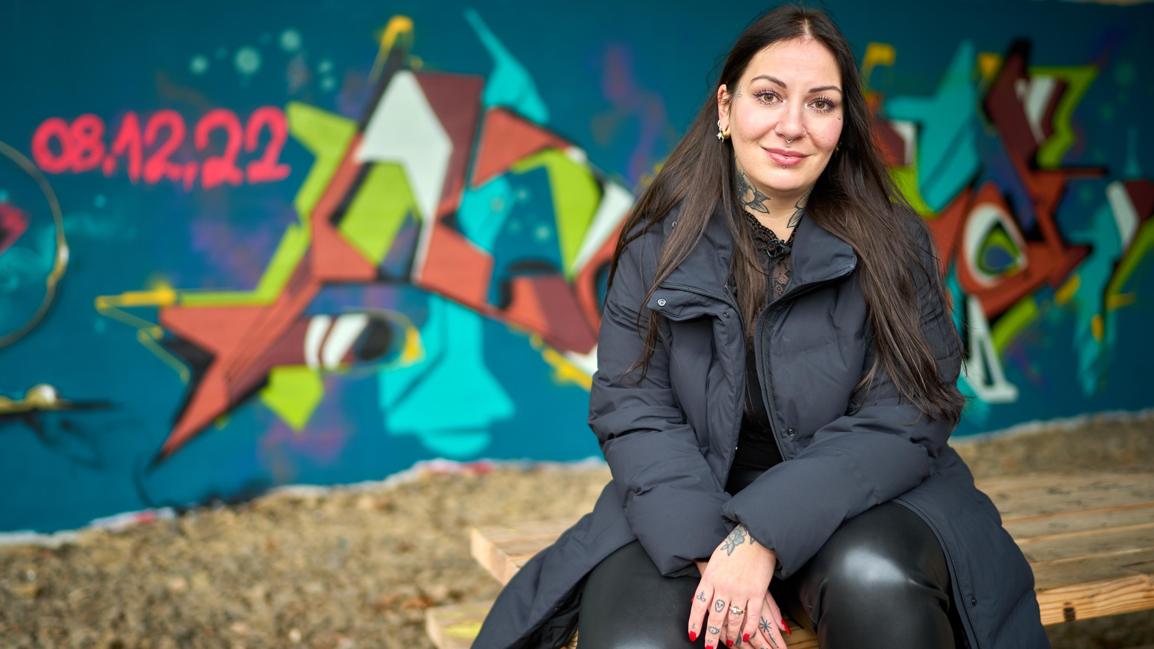 Streetworkerin Amelie Sophie Preyss sitzt auf einem Holztisch vor einer Graffiti-Wand. Sie trägt schwarze Kleidung, Tattoos an Hals und Händen, Piercings und hat rote Fingernägel.