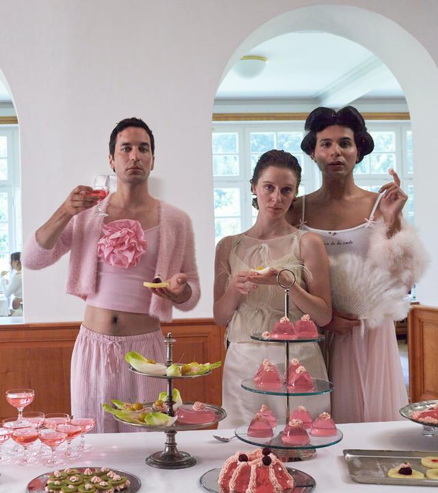 "Unterwegs im Namen der Kaiserin": Karlheinz (Thomas Hauser), Romy (Julia Windischbauer) und Magda Gustav (Benjamin Radjaipour) stehen vor einem Buffet, auf dem kleine Häppchen und Getränke in Gläsern stehen. Alles ist in pinken und weißen Farben gehalten.