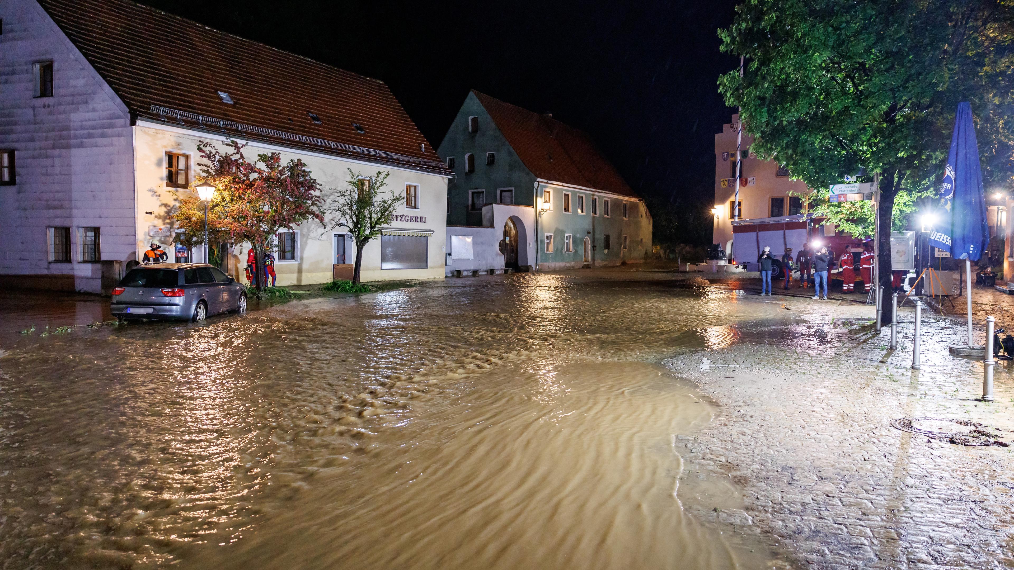 21.05.2024, Bayern, Kastl: Feuerwehrleute arbeiten in den Überflutungen. Wassermassen fließen nach starkem Regen aus dem Fluss Lauterach über den Marktplatz in der Gemeinde Kastl.
