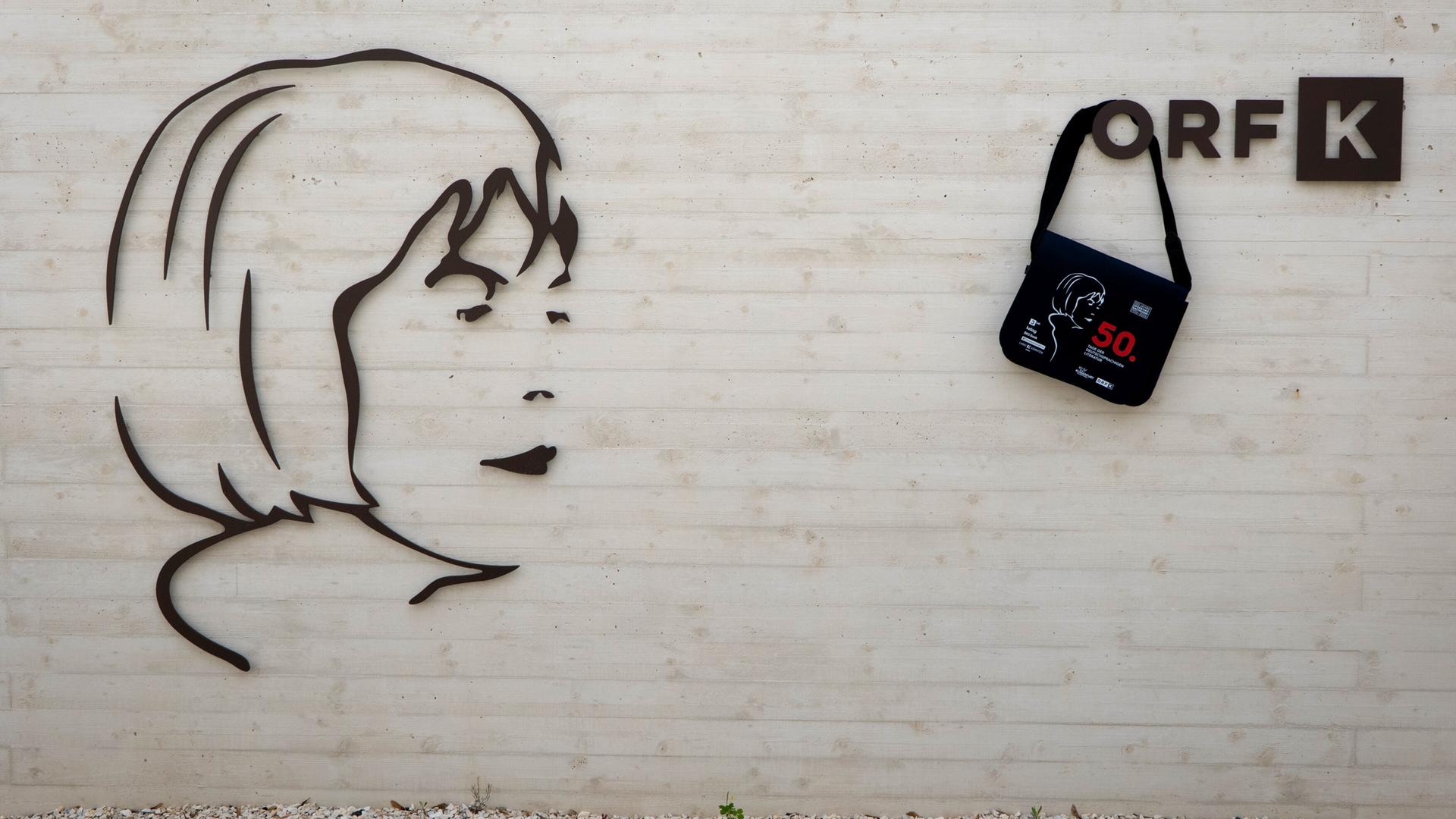 Das Bild zeigt eine Wand des Ingeborg Bachmannparks, die aus hellem Beton besteht. Darauf ist ein stilisiertes, schwarzes Relief einer Gesichtsprofilansicht, das an Ingeborg Bachmann erinnert. Rechts neben dem Relief befindet sich ein schwarzer Tragbeutel, der mit einer grauen Zeichnung eines Gesichts und roten Texten geschmückt ist. Darunter steht die Zahl "50". Über dem Relief und der Tasche ist das Logo „ORF K“ in einem schlichten, modernen Design angebracht. Der Boden vor der Wand besteht aus kleinen, hellen Steinen und es sind einige grüne Pflanzen zu erkennen.