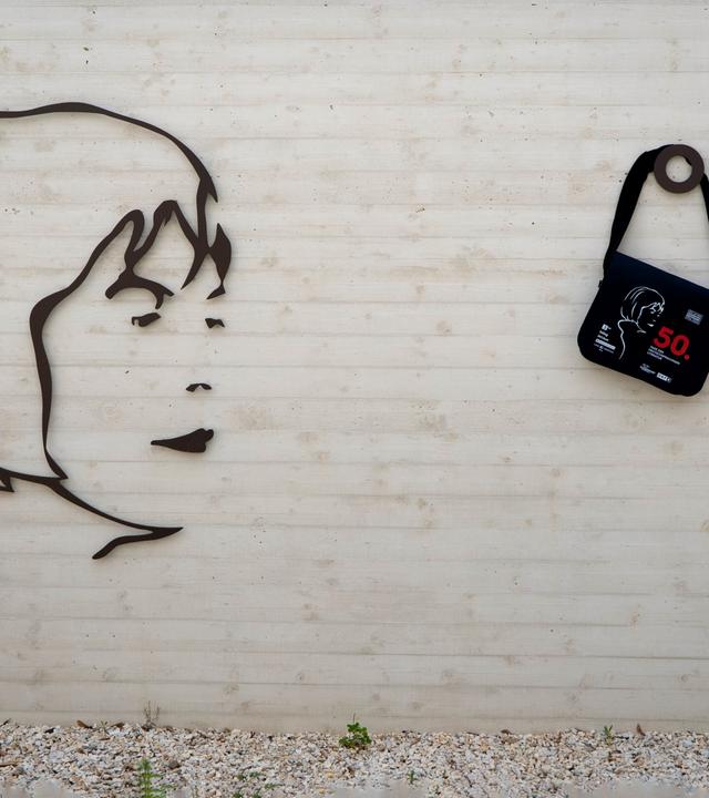 Das Bild zeigt eine Wand des Ingeborg Bachmannparks, die aus hellem Beton besteht. Darauf ist ein stilisiertes, schwarzes Relief einer Gesichtsprofilansicht, das an Ingeborg Bachmann erinnert. Rechts neben dem Relief befindet sich ein schwarzer Tragbeutel, der mit einer grauen Zeichnung eines Gesichts und roten Texten geschmückt ist. Darunter steht die Zahl "50". Über dem Relief und der Tasche ist das Logo „ORF K“ in einem schlichten, modernen Design angebracht. Der Boden vor der Wand besteht aus kleinen, hellen Steinen und es sind einige grüne Pflanzen zu erkennen.