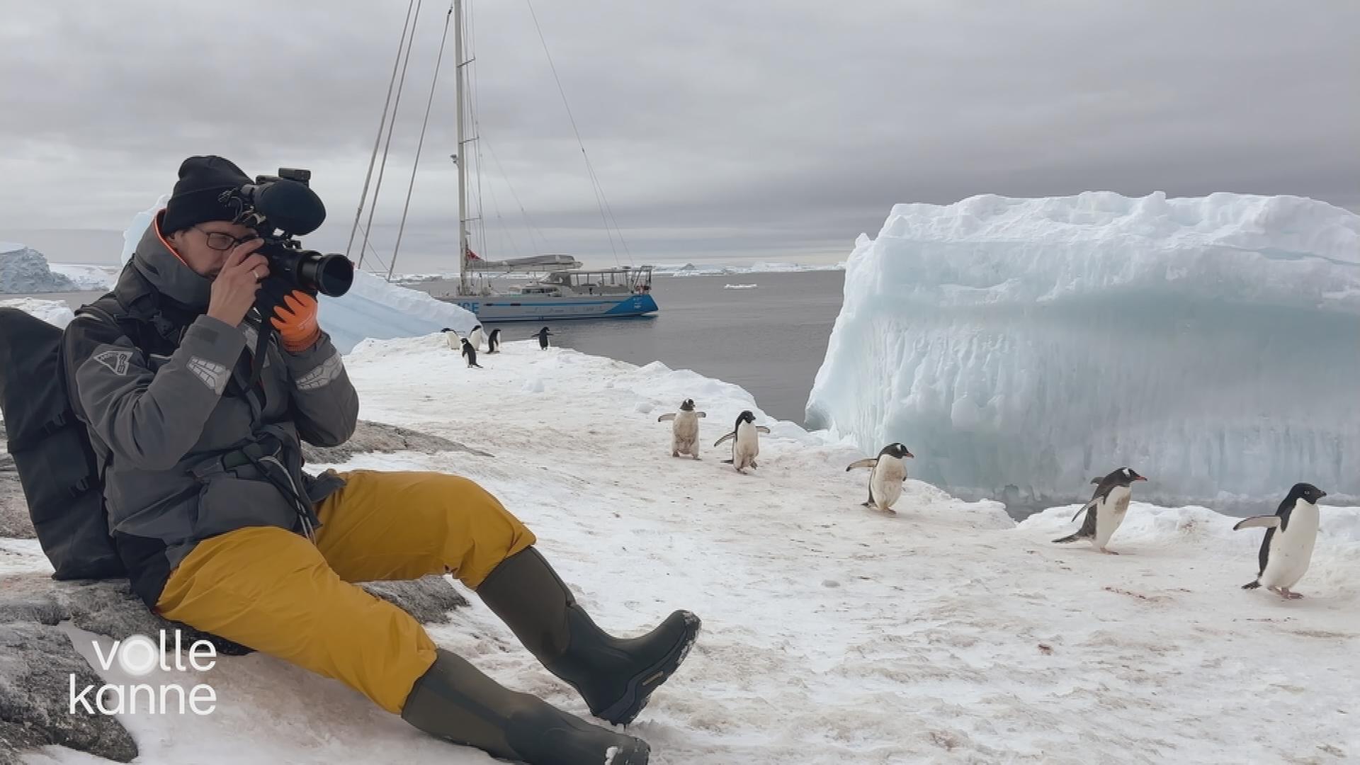 Mann sitzt mit Kamera auf dem Eis. Im Hintergrund sind Pinguine und ein Segelboot in der Antarktis-Landschaft zu sehen.