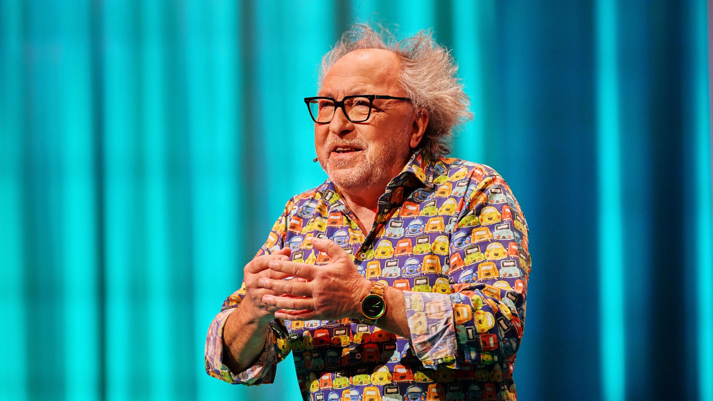 Urban Priol ist in der Mitte des Bildes zu sehen. Er hebt seine Hände vor seine Brust. Sein Hemd ist bunt. Urban Priol trägt eine Brille. Der Hintergrund ist türkis beleuchtet. Am linken Handgelenk trägt er eine Uhr.