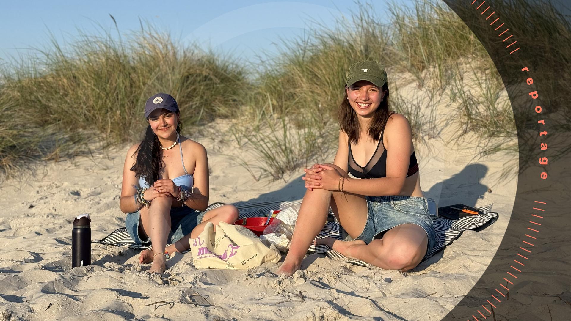 Zwei junge Frauen sitzen beim Picknick in den Dünen und schauen lächelnd in die Kamera.