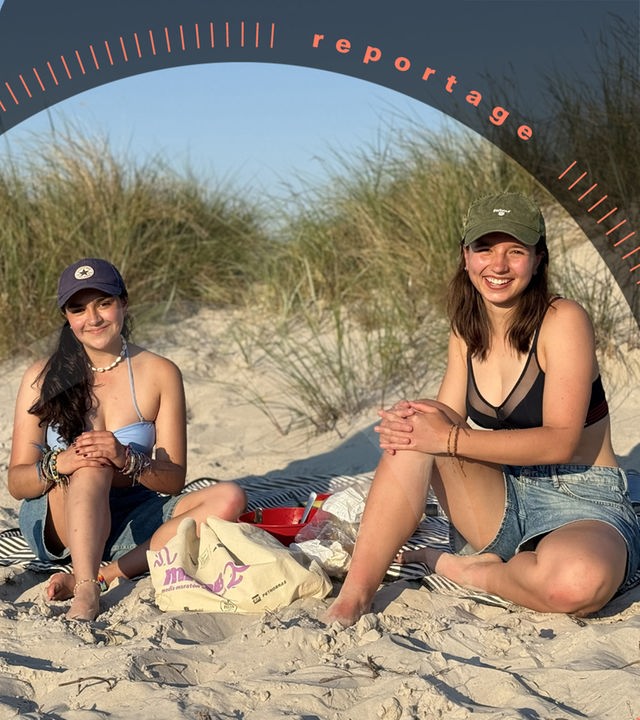 Zwei junge Frauen sitzen beim Picknick in den Dünen und schauen lächelnd in die Kamera.