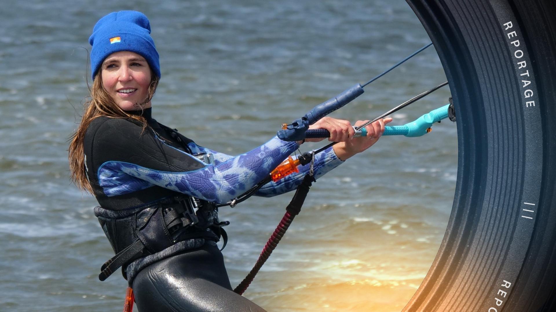 Eine junge Frau im Neoprenanzug und mit Mütze ist im Wasser beim Kitesurfen und hält sich an einem Griff fest. Sie lächelt.