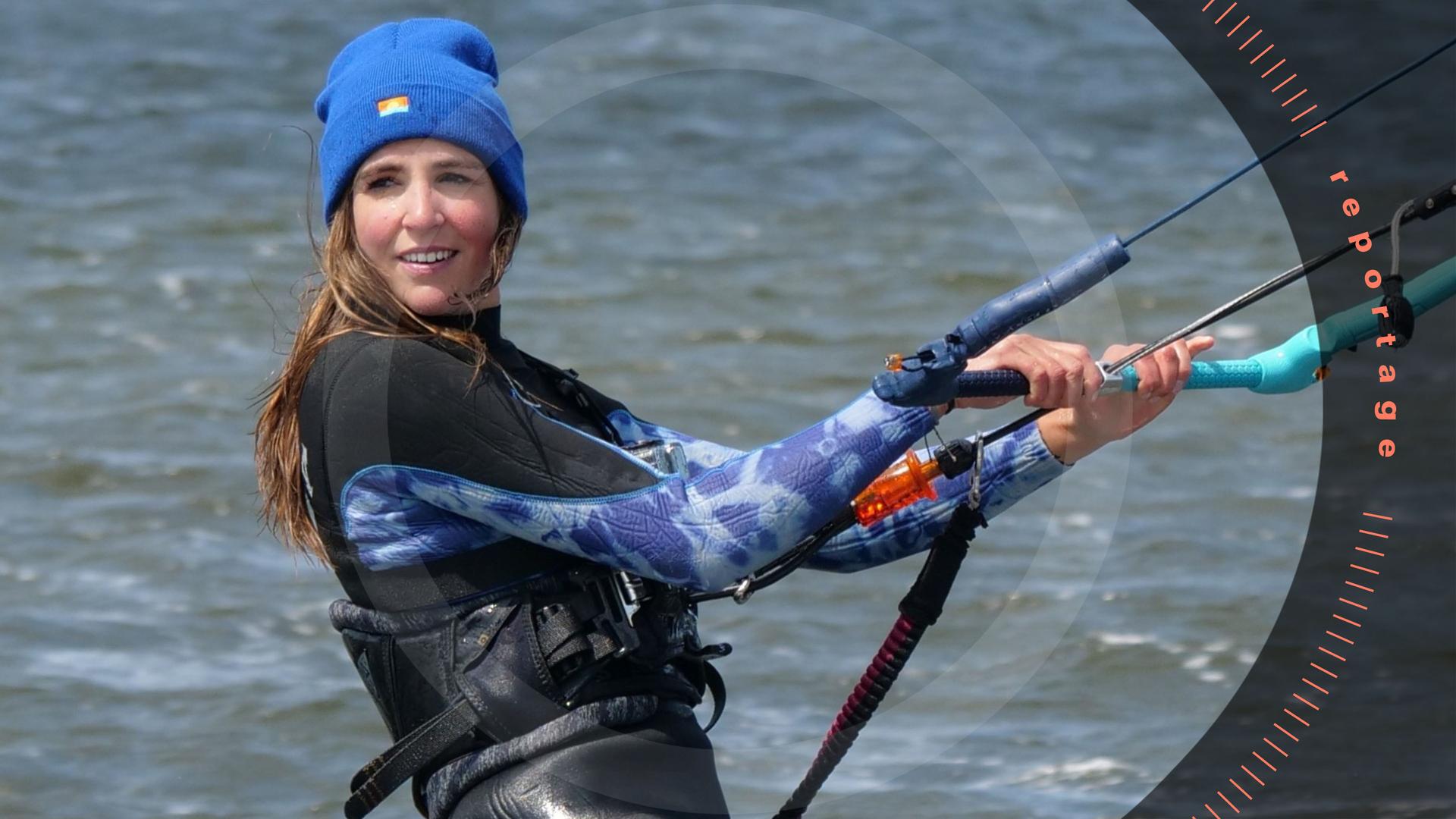 Eine junge Frau im Neoprenanzug und mit Mütze ist im Wasser beim Kitesurfen und hält sich an einem Griff fest. Sie lächelt.