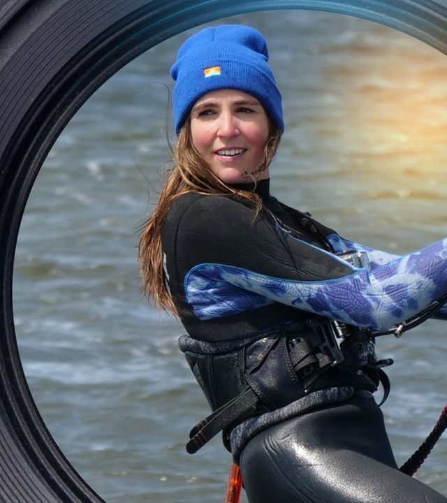 Eine junge Frau im Neoprenanzug und mit Mütze ist im Wasser beim Kitesurfen und hält sich an einem Griff fest. Sie lächelt.