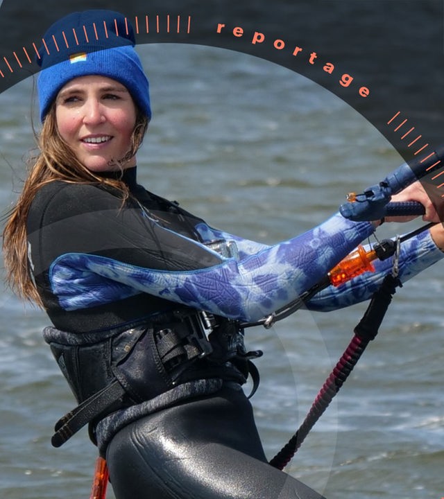 Eine junge Frau im Neoprenanzug und mit Mütze ist im Wasser beim Kitesurfen und hält sich an einem Griff fest. Sie lächelt.