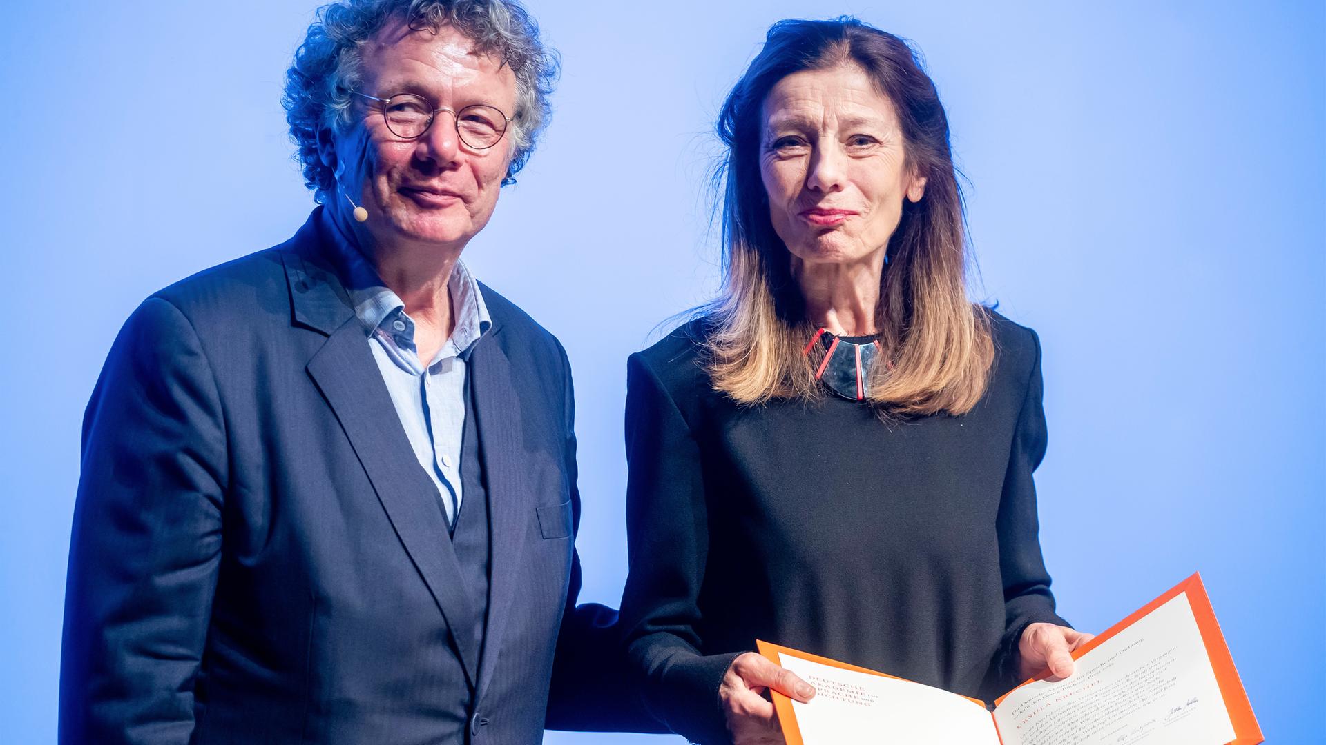 Eine Frau mit langen Haaren hält eine Kladde in den Händen, die sie geöffnet präsentiert. Neben ihr steht ein Mann.