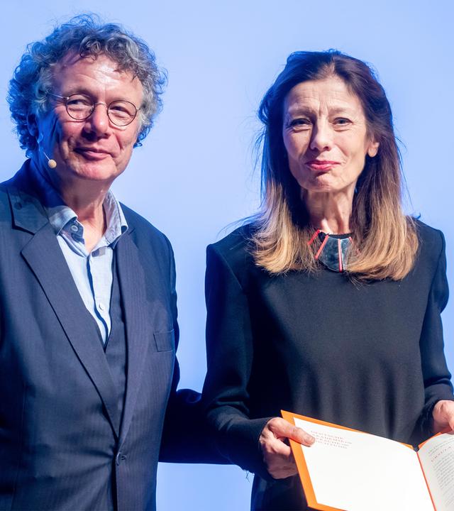 Eine Frau mit langen Haaren hält eine Kladde in den Händen, die sie geöffnet präsentiert. Neben ihr steht ein Mann.