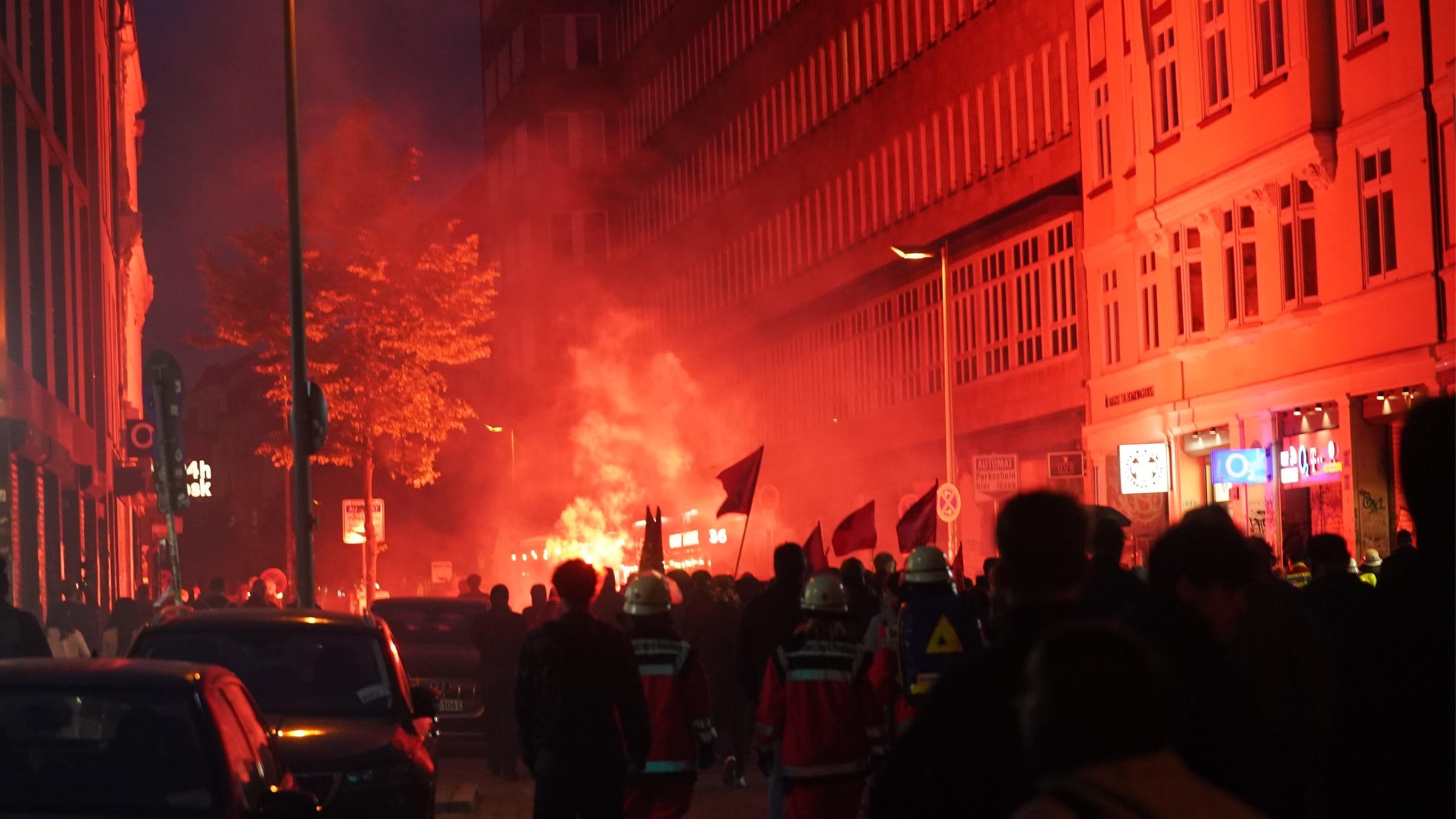 Demonstranten in Hamburg brennen nachts rote Feuerwerkskörper ab. Die Häuserfassade im Hintergrund leuchtet rot. Im Vordergrund eine größere Masse an Demonstranten.
