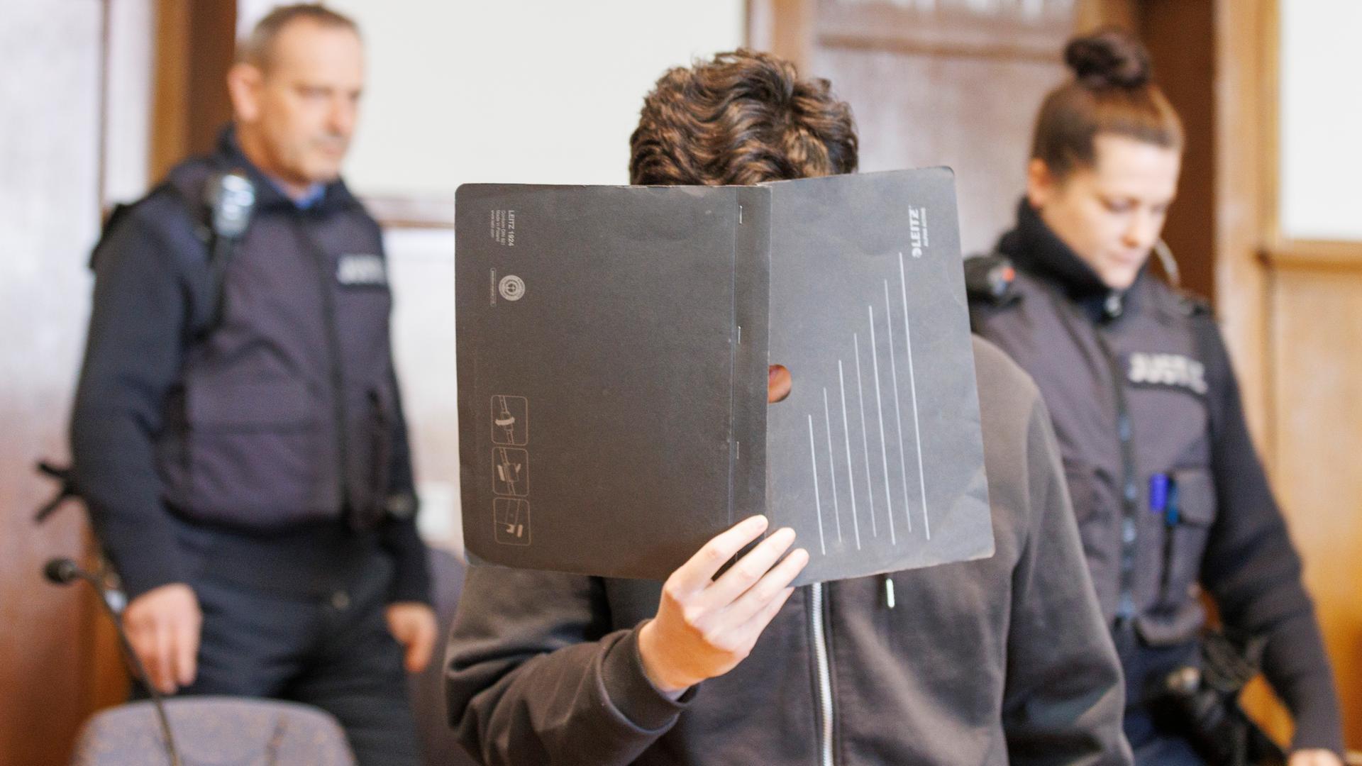 Der Angeklagte ist im Gerichtssaal zu sehen, er verdeckt sein Gesicht. Im Hintergrund sind zwei Polizist*innen zu sehen.
