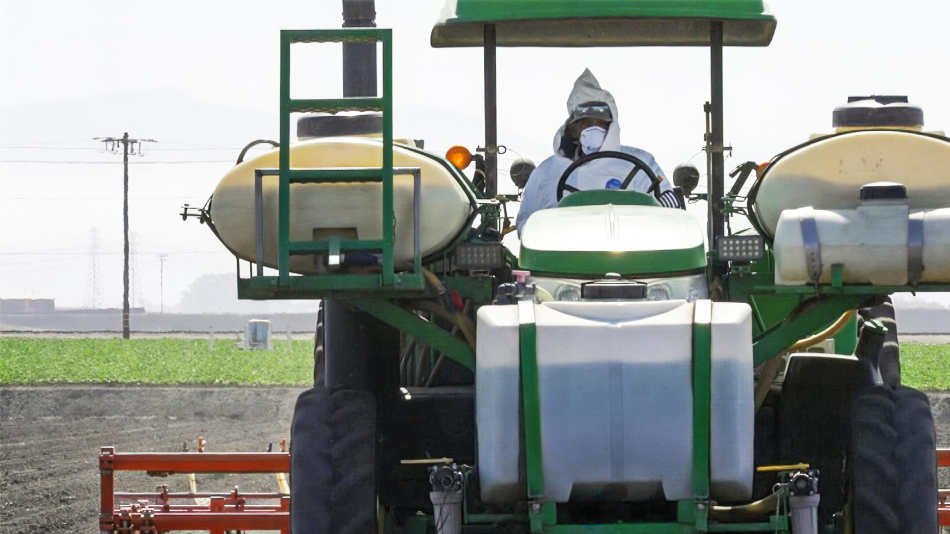 Ein Mann in weißer Schutzkleidung und mit Atemmaske fährt einen Traktor, an dem verschiedene Kunststofftanks befestigt sind.