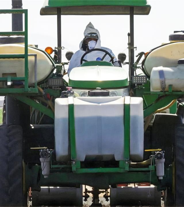 Ein Mann in weißer Schutzkleidung und mit Atemmaske fährt einen Traktor, an dem verschiedene Kunststofftanks befestigt sind.