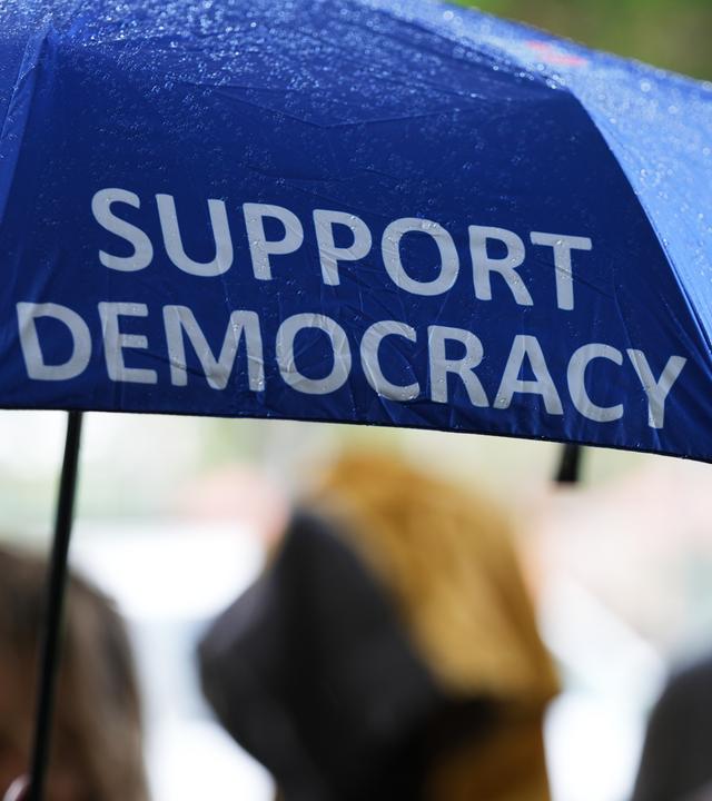 27.10.2026, USA, Coral Gables: Eine Person trägt vor einem Wahllokal einen Regenschirm mit der Aufschrift "Support Democracy" und "No Dictators" während die vorzeitige Stimmabgabe für die Zwischenwahlen 2026 stattfindet.