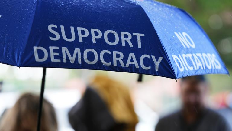 27.10.2026, USA, Coral Gables: Eine Person trägt vor einem Wahllokal einen Regenschirm mit der Aufschrift "Support Democracy" und "No Dictators" während die vorzeitige Stimmabgabe für die Zwischenwahlen 2026 stattfindet.