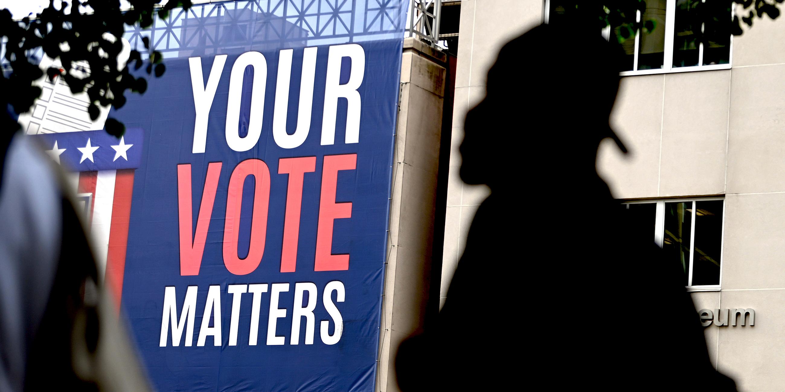 Ein Mann geht an einem Banner vorbei der mit "Your Vote Counts" Passanten erinnert, wählen zu gehen. 