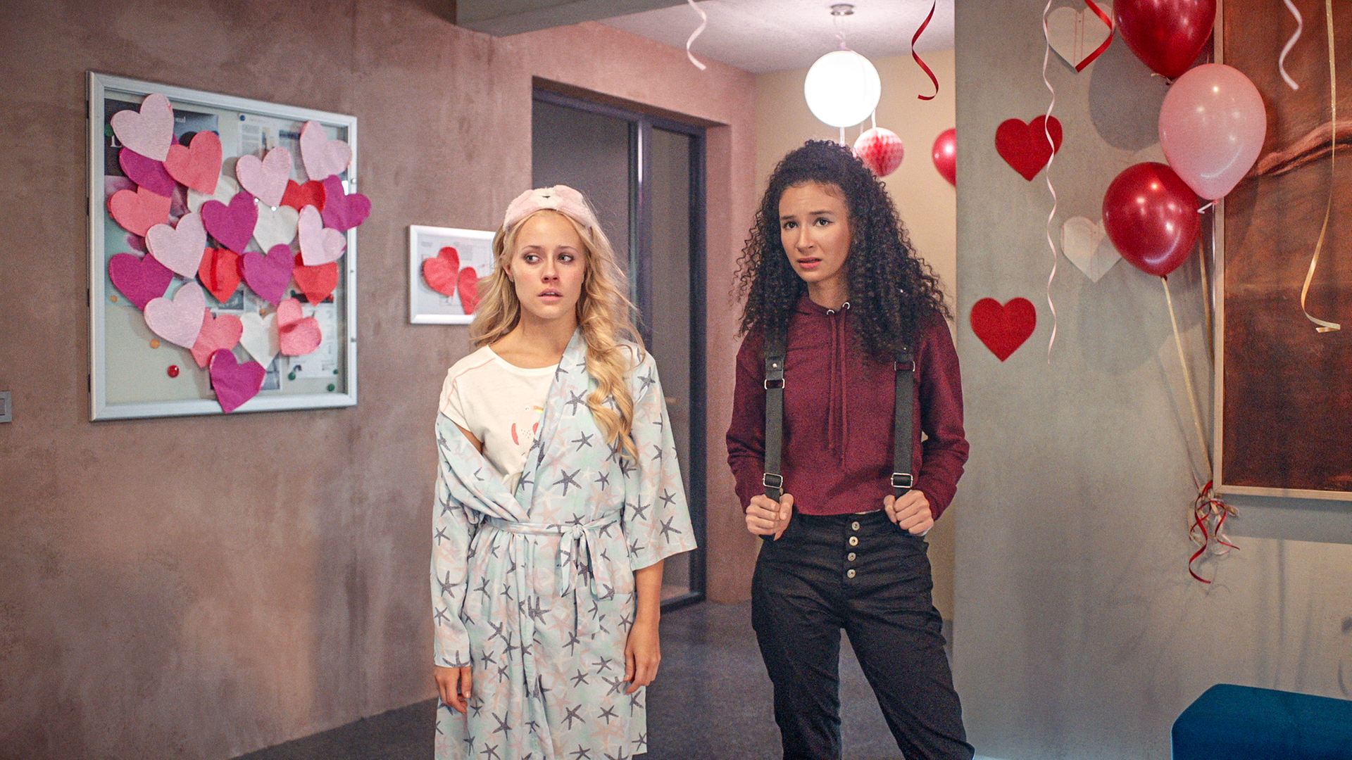 Lena (links) und Ines (rechts) stehen im Flur, der für Valentinstag mit vielen Herzen geschmückt ist. Lena trägt ihren Schlafanzug und einen Bademantel, sie sieht aus als hätte sie viel geweint. Ines sieht besorgt aus.