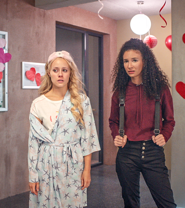 Lena (links) und Ines (rechts) stehen im Flur, der für Valentinstag mit vielen Herzen geschmückt ist. Lena trägt ihren Schlafanzug und einen Bademantel, sie sieht aus als hätte sie viel geweint. Ines sieht besorgt aus.