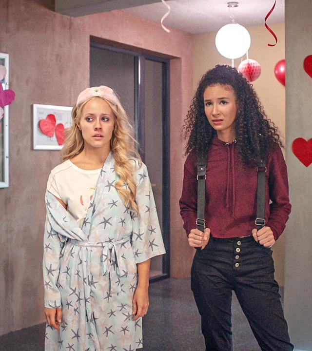 Lena und Ines stehen im Flur, der für Valentinstag mit vielen Herzen geschmückt ist. Lena trägt ihren Schlafanzug und einen Bademantel, sie sieht aus als hätte sie viel geweint. Ines sieht besorgt aus.