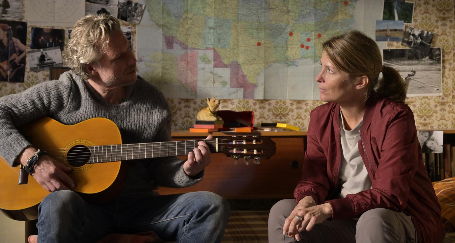 "Nächste Ausfahrt Glück - Beste Freundinnen": Juri (Dirk Borchardt) und Katharina (Valerie Niehaus) sitzen sich in Juris Jugendzimmer auf dem Bett gegenüber. Juri spielt auf einer Gitarre. Im Hintergrund an der Zimmerwand alte Fotos und eine Landkarte der USA.