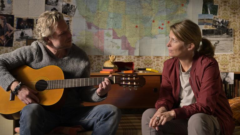 "Nächste Ausfahrt Glück - Beste Freundinnen": Juri (Dirk Borchardt) und Katharina (Valerie Niehaus) sitzen sich in Juris Jugendzimmer auf dem Bett gegenüber. Juri spielt auf einer Gitarre. Im Hintergrund an der Zimmerwand alte Fotos und eine Landkarte der USA.