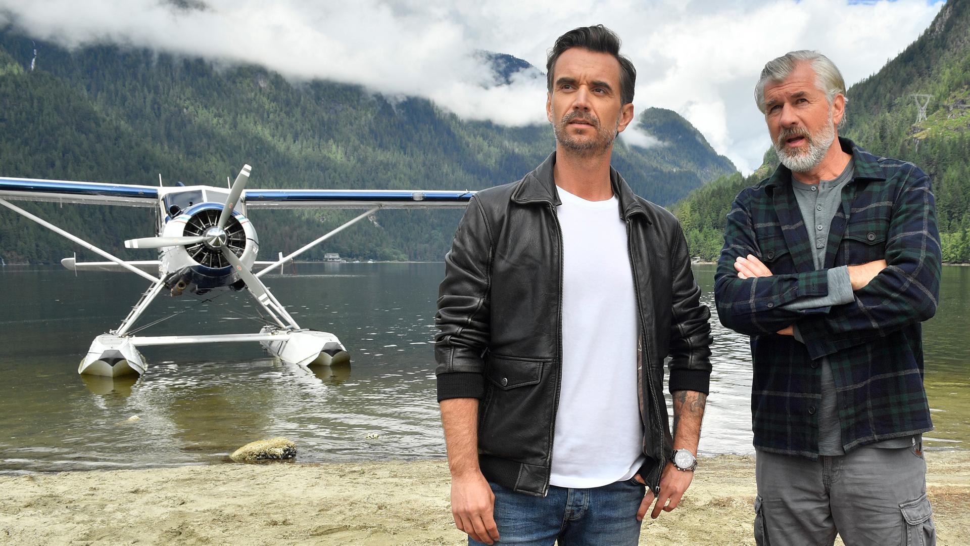 Max Parger (Florian Silbereisen) und Martin Grimm (Daniel Morgenroth) stehen am Ufer eines Sees nebeneinander. Martin hat die Arme vor der Brust verschränkt und redet mit Max. Im Hintergrund steht ein Wasserflugzeug.