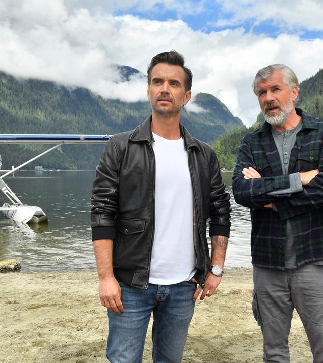 Max Parger (Florian Silbereisen) und Martin Grimm (Daniel Morgenroth) stehen am Ufer eines Sees nebeneinander. Martin hat die Arme vor der Brust verschränkt und redet mit Max. Im Hintergrund steht ein Wasserflugzeug.