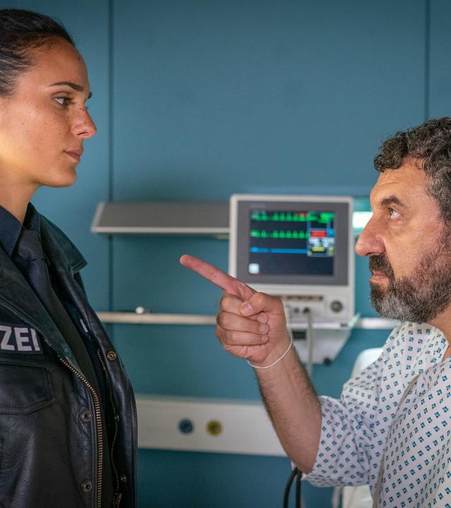 "Notruf Hafenkante - Vatertag": Tamer Aslan (Raymond Tarabay) sitzt auf seinem Bett in einem Krankenzimmer des Elbkrankenhause. Er weist mit seinem rechten Zeigefinger warnend auf die Polizistin Pinar Aslan (Aybi Era), die vor ihm steht. Beide sehen sich ernst in die Augen.