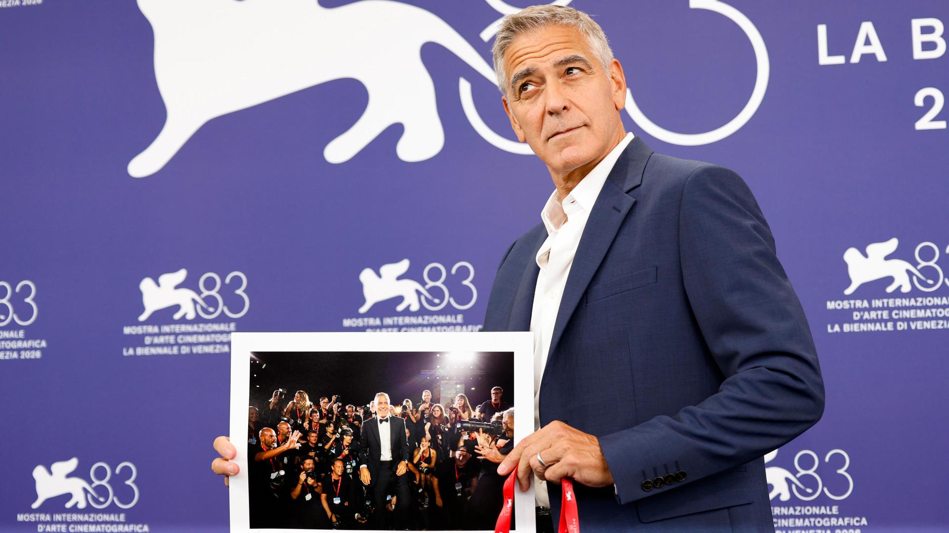Ein Mann im Anzug posiert vor der Fotowand des Filmfestivals Venedig mit einem Bild von sich selbst.