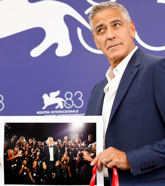 Ein Mann im Anzug posiert vor der Fotowand des Filmfestivals Venedig mit einem Bild von sich selbst.