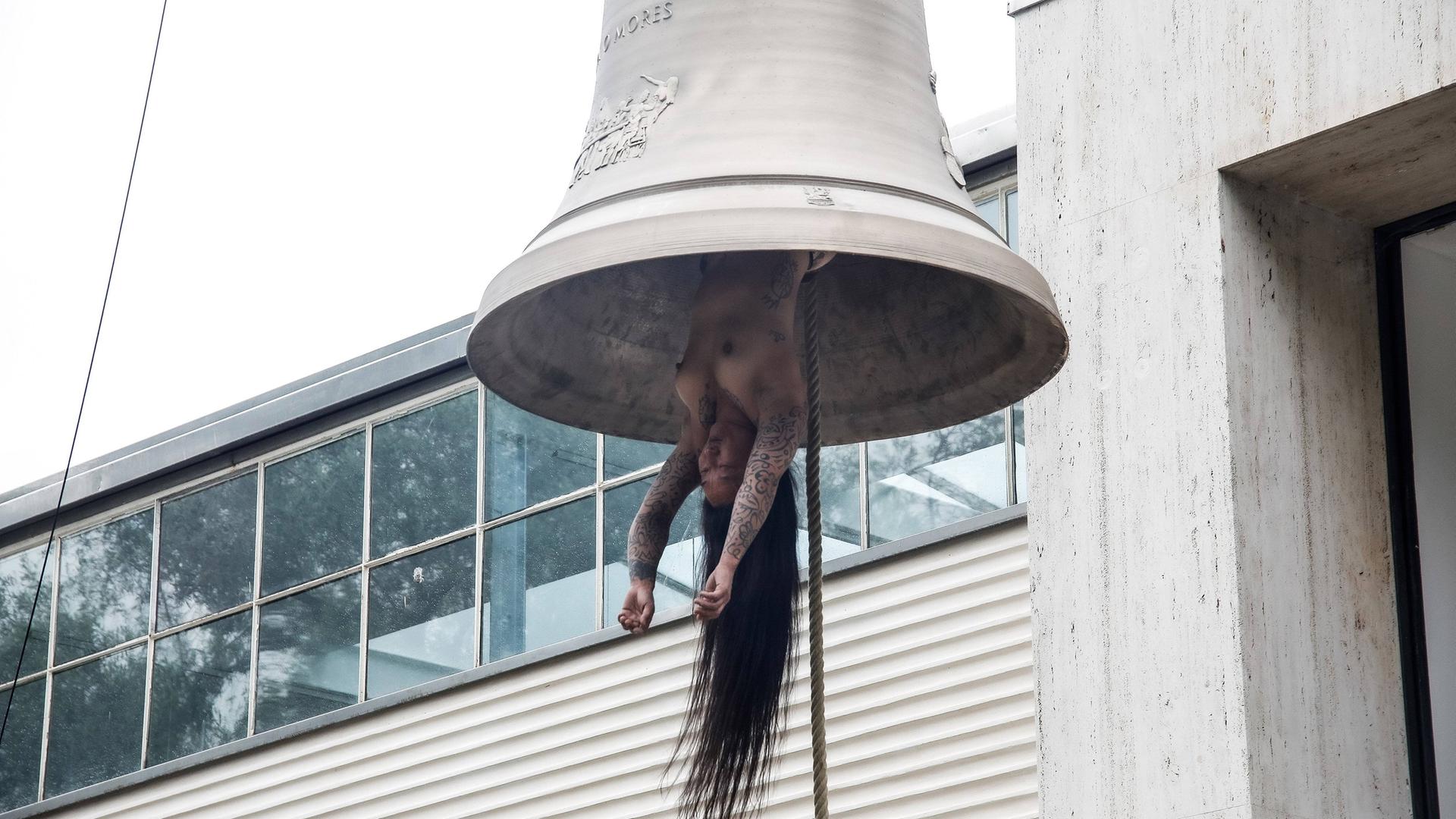 Eine nackte tätowierte Frau mit langen Haaren hängt wie ein Schlägel in einer Glocke.