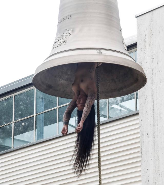 Eine nackte tätowierte Frau mit langen Haaren hängt wie ein Schlägel in einer Glocke.