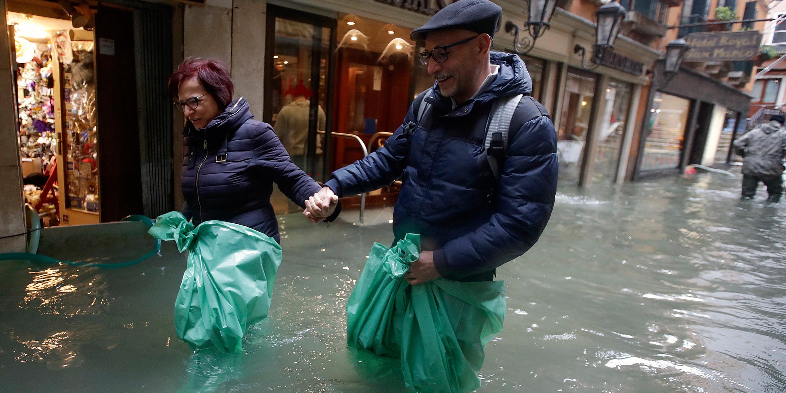 Ein Paar watet knietief in grünen Müllsäcken im Hochwasser einer überfluteten Straße. Am 15.11.2019 in Venedig, Italien.