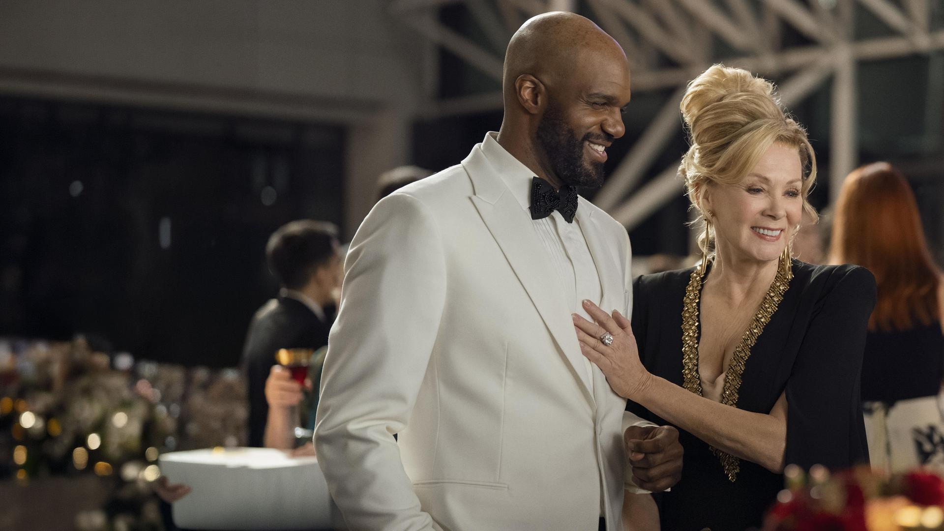 "Hacks - Verändere die Welt mit Lachern": Deborah (Jean Smart) steht im schicken Abendkleid neben dem mit weißen Smoking festlich gekleidetem Marcus (Carl Clemons-Hopkins). Deborah hat ihre Hand auf dessen Brust gelegt und beide lachen glücklich und gelöst. Im Hintergrund befinden sich weitere Personen (Komparsen).