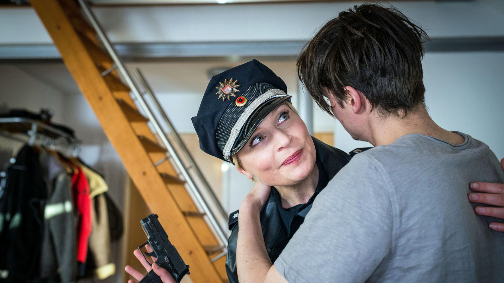 "Notruf Hafenkante - Verbotene Liebe": Die Polizistin Melanie Hansen (Sanna Englund) steht in einem Wohnraum dicht neben Fabian Westphal (Gordon Kämmerer), der seine Arme um ihren Hals gelegt hat. Melanie lächelt Fabian verhalten an. In ihrer rechten Hand hat sie ihre Pistole, die sie sichernd seitlich ihres Körpers hält.