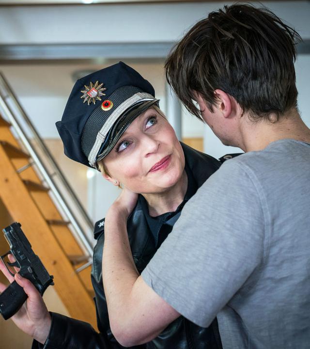 "Notruf Hafenkante - Verbotene Liebe": Die Polizistin Melanie Hansen (Sanna Englund) steht in einem Wohnraum dicht neben Fabian Westphal (Gordon Kämmerer), der seine Arme um ihren Hals gelegt hat. Melanie lächelt Fabian verhalten an. In ihrer rechten Hand hat sie ihre Pistole, die sie sichernd seitlich ihres Körpers hält.