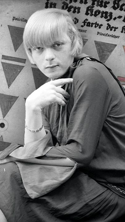 Transfrau Liddy Bacroff im Vordergrund. Im Hintergrund links Gefangene im Konzentrationslager Sachsenhausen; rechts eine Tafel mit KZ-Kennzeichen.